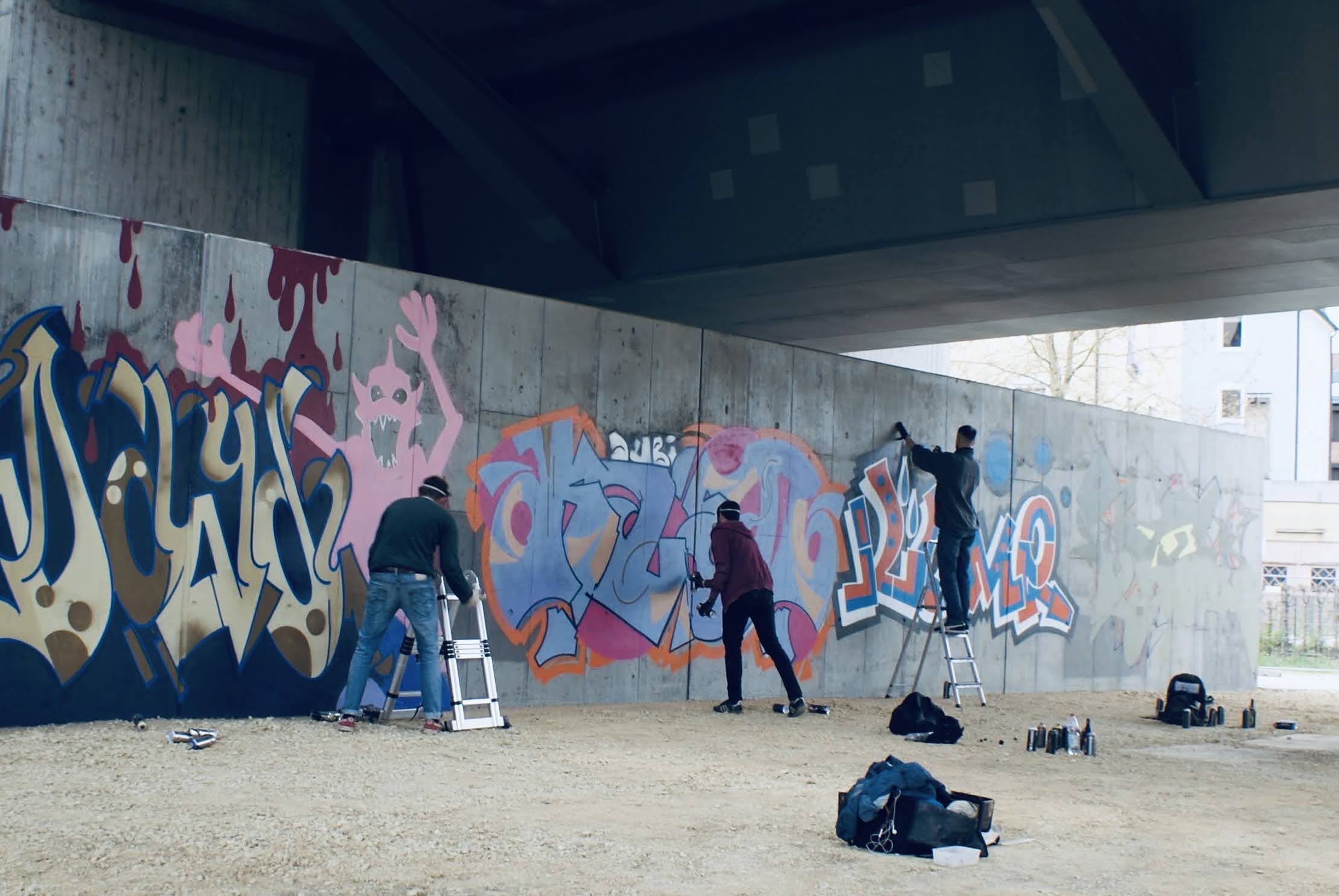Graffiti an der HWS Wand. Drei Graffiti Künstler besprühen die graue Betonwand mit bunten Schriftzügen. Die Grundform eines rosa Monsters ist in der linken Bildhälfte zu erkennen.