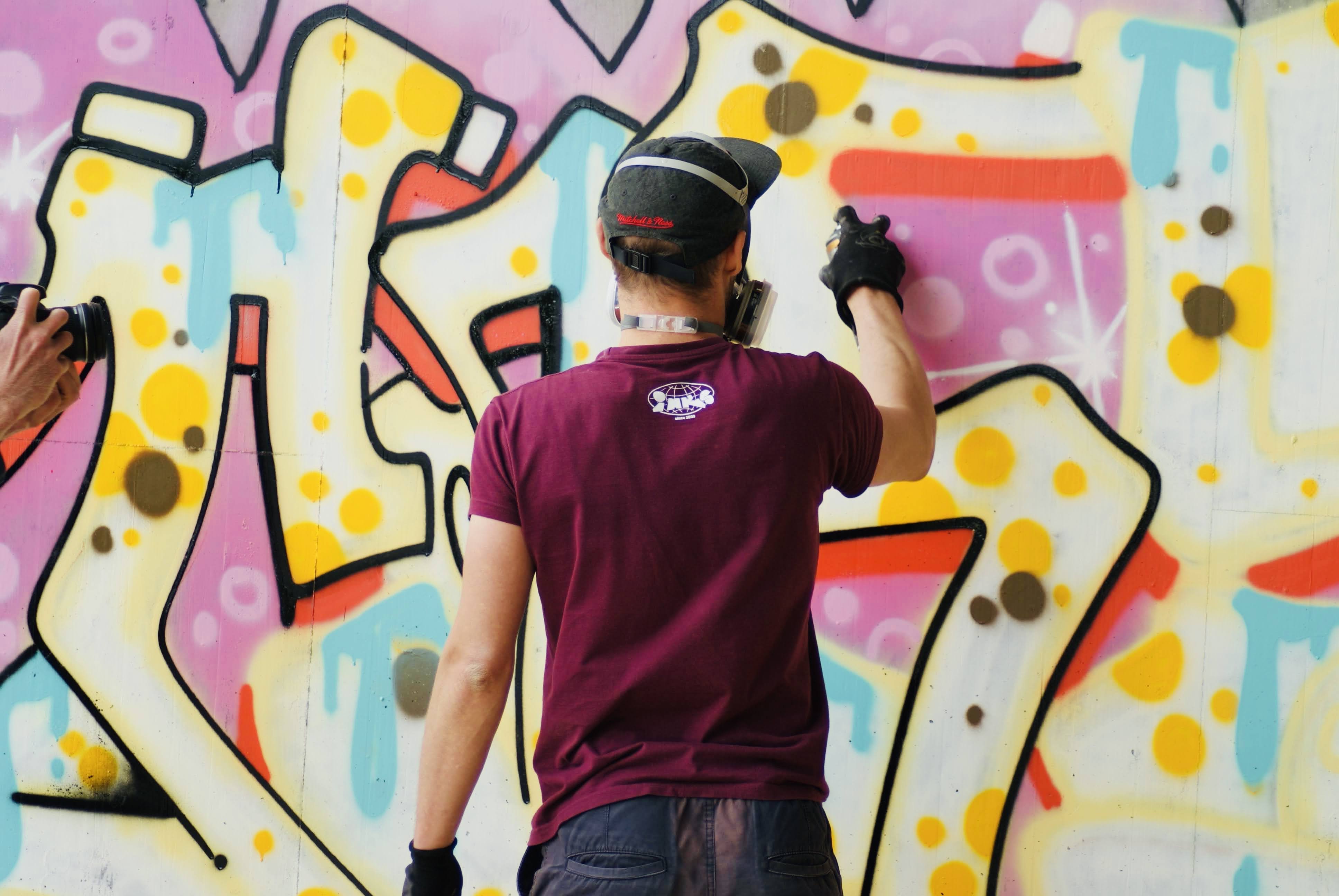 Graffiti an der HWS Wand. Ein Graffiti Künstler in der Mitte des Bildes mit dem Rücken zur Kamera im dunkelroten Shirt und Baseballkappe sprüht mit frischen Pastellfarben einen Schriftzug an die Mauer.